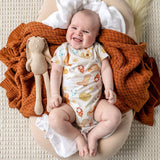 A smiling baby lies on a beige cushion wrapped in a chunky rust-orange knit blanket, wearing a cream bodysuit with a colorful space-themed print. A beige plush bunny toy rests beside the baby.