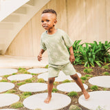 Child in a green outfit with palm tree pattern standing on white stones outdoors.