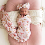 A sleeping newborn baby is swaddled in a light pink western-print swaddle blanket and wearing a matching bow headband, resting on a pink maternity pillow with a white knit blanket.