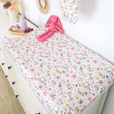 A white changing table covered with a white fabric cover printed with a colorful watercolor-style floral pattern. A pink plush bunny toy and a small tray of baby accessories rest on the table, set in a bright nursery-like room with white drawers and wall decor.