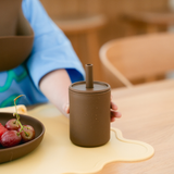 Person holding a brown ceramic cup with a straw on a wooden table.