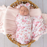 A sleeping newborn baby is swaddled in a pink and white flamingo floral blanket and wrapped in a matching pink knitted swaddle, resting in a wicker basket with a pink blanket.
