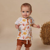 A toddler stands against a warm beige studio backdrop wearing a white short-sleeve top with a western-themed print of cowboy boots, horseshoes, stars, and blocks, paired with brown shorts. A bale of hay sits to the right.