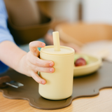 Hand holding a yellow cup with a straw on a wooden table.