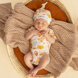 A sleeping newborn baby wearing a matching cream short-sleeve bodysuit and knit beanie hat with a colorful animal print, resting in a woven basket bassinet with a brown cushion and a chunky knit beige blanket.