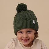 Child wearing a green knit beanie with a pom-pom on a beige background