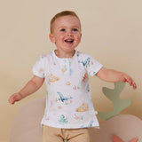 A smiling toddler wearing a white short-sleeve t-shirt with a colorful underwater sea-creature print, standing in front of a beige background.