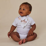 A baby sitting cross-legged wearing a white short-sleeve bodysuit and a white bandana bib with a horse and donkey print, set against a solid beige background.