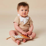 A baby sitting cross-legged on a beige studio background, wearing a beige short-sleeve romper and a white bib with a small animal illustration. The baby is holding a wooden xylophone toy.
