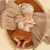 A sleeping newborn baby wearing a matching cream and orange bakery-themed outfit with a hat, resting in a woven basket bassinet with a knit blanket.
