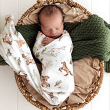 A sleeping newborn baby swaddled in a white wrap blanket with a brown and green desert safari print, resting inside a woven wicker basket lined with beige fabric and a dark green knit blanket.