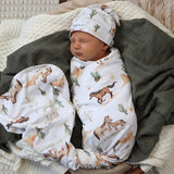 A sleeping newborn baby swaddled in a white blanket with a brown and green western print, wearing a matching hat, resting on a dark olive green blanket and cream knit pillows.
