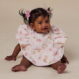 A baby wearing a ruffled bib with a pink cowboy boot and floral print, sitting on a beige studio background.