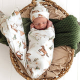 A sleeping newborn baby is swaddled in a white muslin blanket with a brown and green safari print, matching with a hat. The baby is nestled inside a round woven basket lined with tan fabric and a textured dark green blanket.