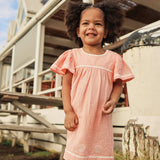A young child with curly hair wearing a light pink gingham nightgown with white lace trim, standing outdoors in front of a rustic white wooden structure.