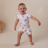 A smiling baby wearing a white short-sleeve romper with a teddy bear print and beige trim, standing barefoot against a warm beige studio background.
