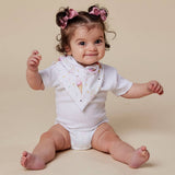 A baby girl sitting on a beige studio background, wearing a white short-sleeve bodysuit with a pink and yellow ice cream cone print bib, and pink hair bows.