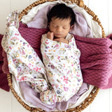 A sleeping newborn baby swaddled in a white floral blanket, resting inside a woven wicker basket lined with lavender and cream fabric and wrapped in a textured magenta knit blanket.