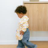 Child wearing a white shirt with yellow patterns and blue jeans walking indoors.