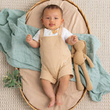 A baby lying on their back in a woven oval bassinet, wearing beige overall shorts over a white t-shirt. A dusty blue-green muslin blanket is draped around them, and a tan plush bunny toy rests beside the baby.