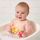 Baby in a bathtub holding two bath toys, one yellow and one red, against a tiled bathroom wall.