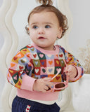 Child wearing a colorful heart-patterned shirt with pink bib and pants, holding sunglasses.