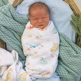 A newborn baby is swaddled in a white muslin blanket with a colorful sea-creature print, lying in a wicker bassinet with a light blue pillow and a green knit blanket.