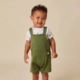 A smiling toddler wearing olive green short overalls over a white short-sleeve t-shirt with a light print, standing against a solid beige background.