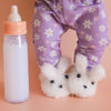 Child wearing purple floral pants with white dog slippers and a baby bottle on a peach background