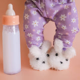 Child wearing purple floral pants with white dog slippers and a baby bottle on a peach background