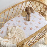 A close-up of a baby bassinet with a white fitted sheet printed with a soft beige teddy bear and bunny pattern, styled with a cream knit blanket and a small plush bunny toy.