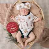 A sleeping newborn baby wearing a white short-sleeve bodysuit and matching bow headband, both printed with teddy bears and Christmas-themed illustrations, resting in a wicker basket with a knit blanket. A red circular sticker reads 'MY FIRST Christmas'.