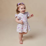 A toddler girl walking barefoot in a white short-sleeve dress with a colorful floral print, featuring purple bows in her hair, set against a solid beige studio background.