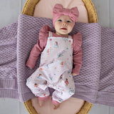 Baby in pink outfit with floral overalls and bow headband lying on a textured blanket.