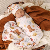 A sleeping newborn baby wrapped in a western-themed swaddle blanket and wearing a matching hat, styled on a cozy blanket with a rust-colored throw.