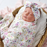 A sleeping newborn baby swaddled in a white muslin blanket with a colorful wildflower print, wearing a matching bow headband, resting in a woven basket.
