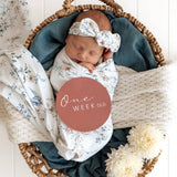 Newborn baby wrapped in a floral blanket with a 'One Week Old' label, surrounded by soft textures and flowers.