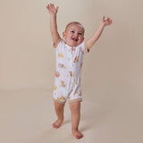 A baby standing barefoot on a warm beige studio background, wearing a sleeveless white footless sleeper with a front zipper and a playful print of teddy bears, bees, and flowers. The baby has their arms raised in the air, surrounded by floating bubbles.