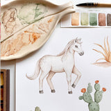 A watercolor painting of a white horse with cacti, shown alongside a decorative ceramic paint palette, a paintbrush, and a small color swatch card on a white background.