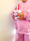 A child in a pink hoodie and matching sweatpants holds a white reusable coffee cup with a red straw and cherry print. The cup has the word 'beigno' printed on it in black text.