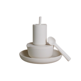 A minimalist white ceramic dinnerware set featuring a shallow bowl with a matching lid, a cylindrical cup with a straw, and a spoon resting on a shallow saucer, all photographed against a seamless white background.