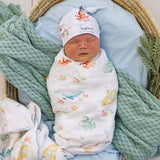 A sleeping newborn baby swaddled in a white muslin blanket with colorful sea-creature prints, wearing a matching white hat with an octopus and the text 'Simply Honey'. The baby is nestled in a wicker bassinet with a light blue cushion, a mint green knit blanket, and greenery.
