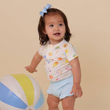 Child wearing a colorful shirt with ice cream cone pattern and light blue shorts, standing next to a beach ball on a beige background.