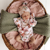 Newborn baby in floral outfit and hat lying on pink blanket in wicker basket with green textured blanket