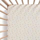 A fitted crib mattress cover with a cream background and a repeating pattern of small gold stars, horseshoes, and letters, shown inside a wooden crib.