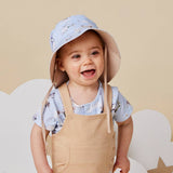 A smiling toddler wearing a light blue sun hat with a beige brim and matching beige overalls, standing against a warm beige background with white cloud shapes.