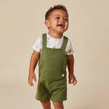 A toddler wearing olive green corduroy overall shorts over a white short-sleeve t-shirt with a subtle floral print, standing against a solid beige background.