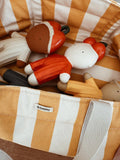 A close-up of a striped fabric storage basket or organizer containing wooden toy figures. The basket has vertical stripes in warm mustard yellow and cream, with a visible brand label reading 'GONMU'.