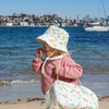 Child on a beach wearing a colorful sun hat and holding a matching bag with a cityscape in the background.