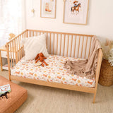 A light natural wood crib or toddler bed styled in a nursery, featuring a cream mattress with a colorful cartoon print, a white pillow, a brown plush toy, and a beige throw blanket.